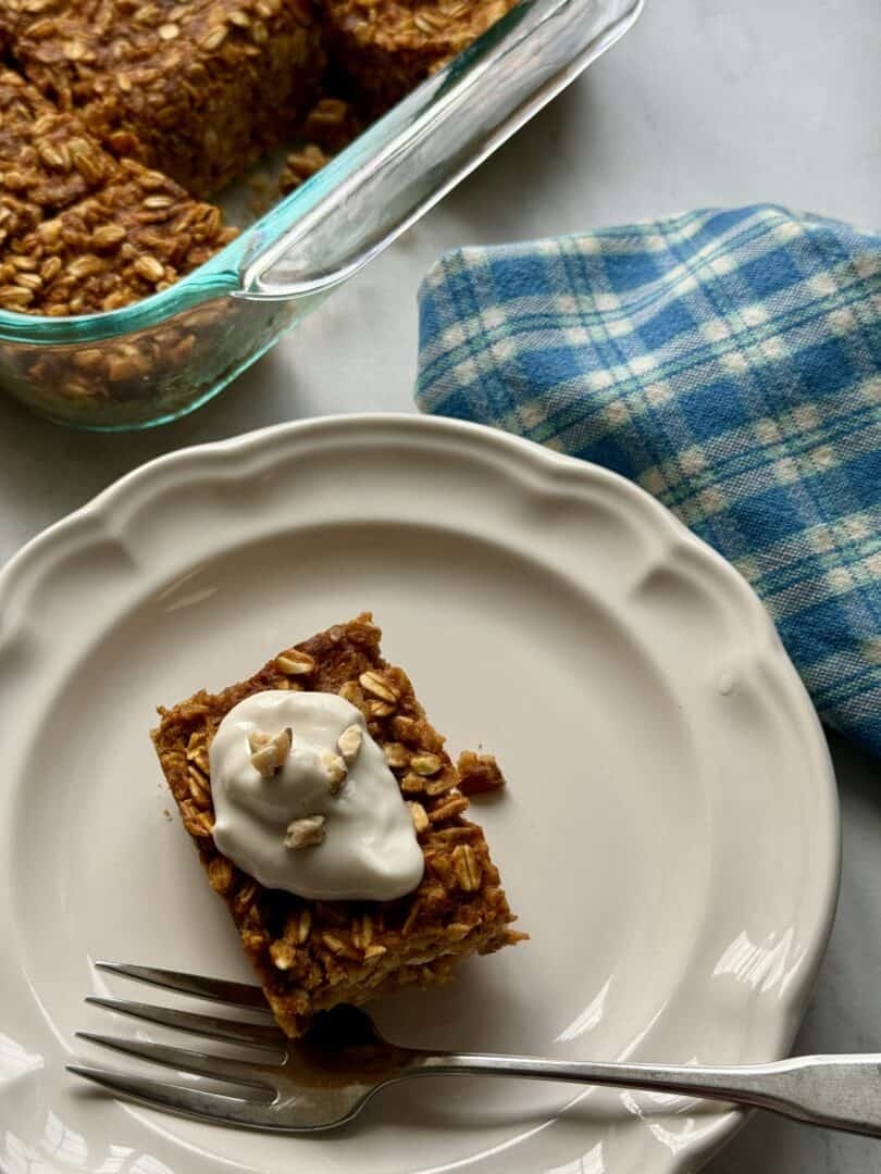 a rectangular piece of vegan pumpkin baked oatmeal on a white plate topped with a dollop of yogurt