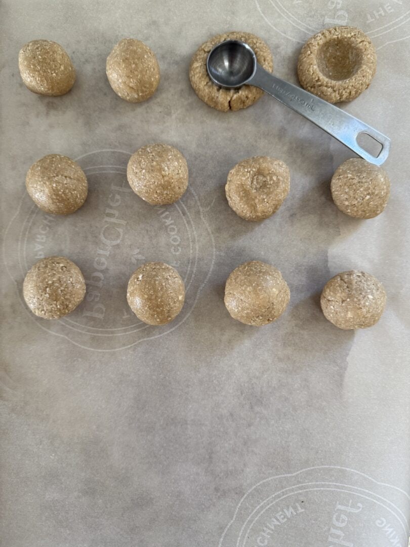 gluten-free thumbprint cookies on a baking sheet with a measuring spoon making an indentation